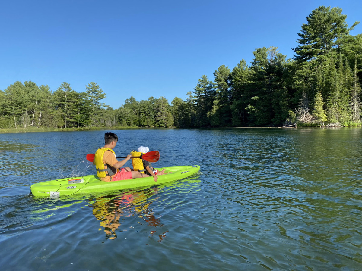 Kayaking on calm forest lake Two people in yellow life jackets paddling a green kayak on a lake surrounded by trees.